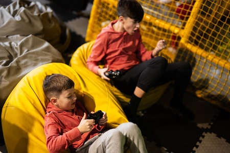 I am winner! Two brothers playing video game console, sitting on yellow pouf in kids play center.