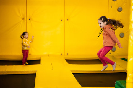 Siblings jumping on trampoline at yellow playground park. Sisters in motion during active entertaiments.の写真素材