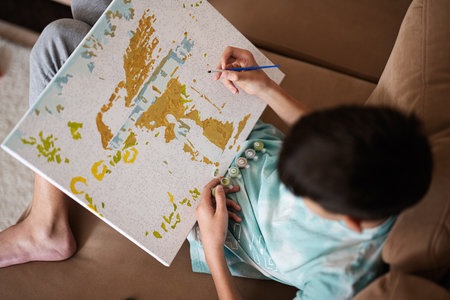 Young male teenager painting picture by numbers on canvas in living room at home. Hobby and leisure concept.の写真素材