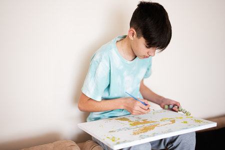 Young male teenager painting picture by numbers on canvas in living room at home. Hobby and leisure...