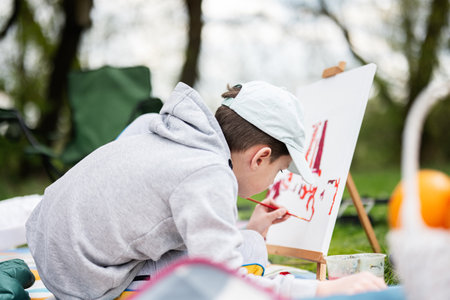 Boy enjoying outdoor on picnic blanket and painting on canvas at garden spring park, relaxation.