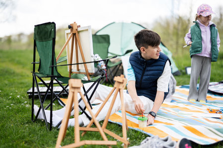 Happy young family, brother and sister having fun and enjoying outdoor on picnic blanket painting ...