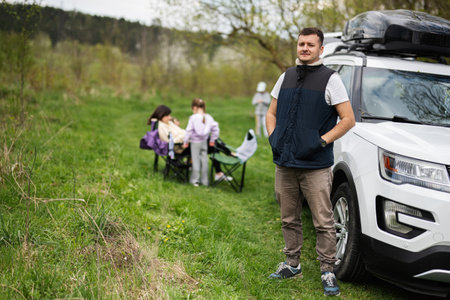 Man in sleeveless vest against suv car with roof rack box, family on picnic.