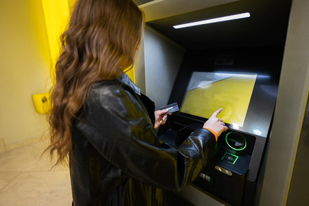 Back of young brunette girl withdrawing money from credit card at yellow ATM.