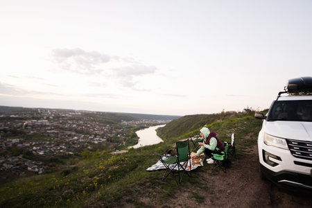 Woman  sit on chair, hiking on vacation, having evening picnic on top of the mountain, looking at beautiful canyon landscape. Car with roof rack box.の写真素材