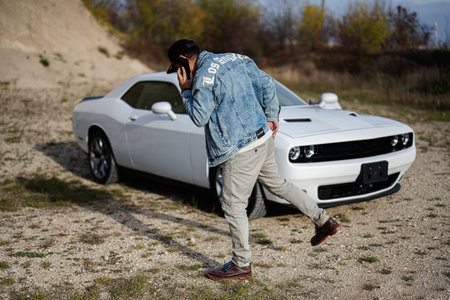 Handsome man in jeans jacket and cap calling on phone while standing near his white muscle car in...