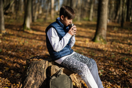 Autumn outdoor portrait of teenager boy sit in stump and drink tea from cup in forest.の写真素材