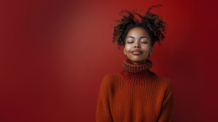 A cheerful young woman in an orange sweater with closed eyes and a smile on her face against a vibrant red background.の素材