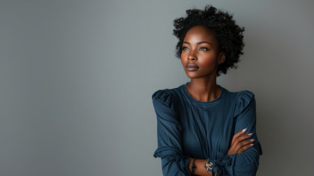 A serious African American woman with natural curly hair and crossed arms in a blue blouse, looking into the distance.の素材