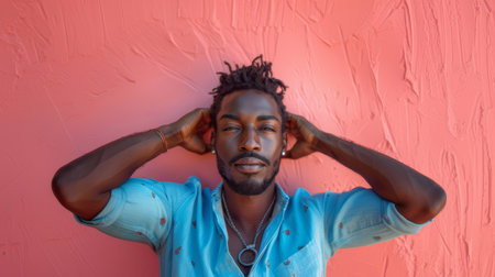 A confident young black man with a stylish look leaning against a pink wall while wearing a blue shirt.の素材