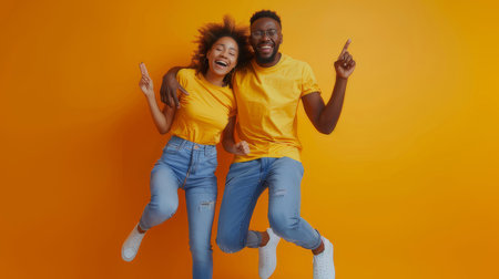Excited African American couple jumping together and celebrating, wearing matching yellow tops and jeans, against a vibrant orange background, showcasing happiness and joy.の素材