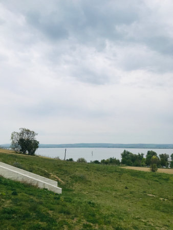Serene landscape featuring a tranquil lake surrounded by lush green hills under a cloudy sky,...
