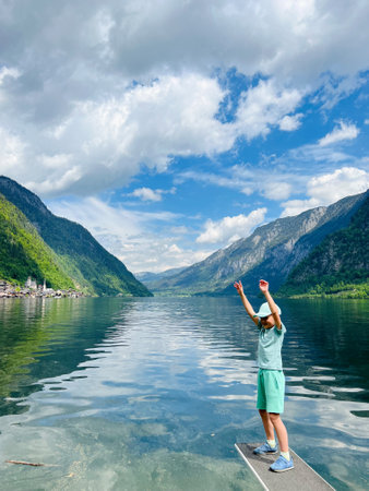 A child joyfully stands at a lake's edge surrounded by majestic mountains and a vibrant sky in Hallstatt, Austria. The scene captures the beauty of nature and the excitement of summer adventures.の写真素材