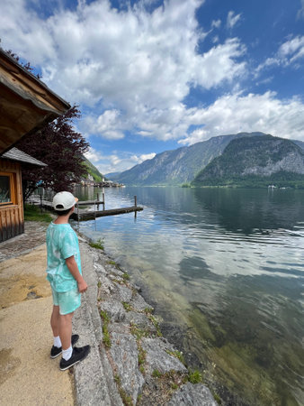 A young boy stands by a serene lake, taking in the mountainous landscape under a partly cloudy sky...