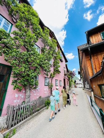 A family walks leisurely down a charming street bordered by pink houses in Hallstatt, Austria.. Lush greenery climbs the walls, adding to the serene and picturesque setting under a bright blue sky.の写真素材