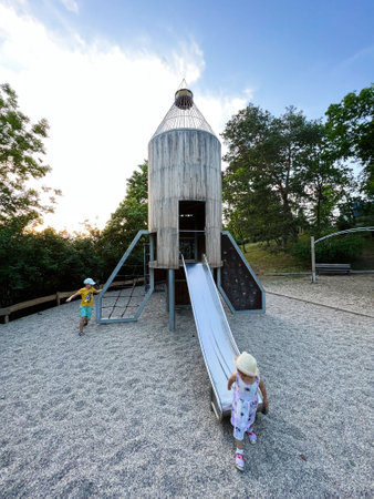 Two young children play happily on a slide in a modern outdoor playground surrounded by trees. The scene captures the joy and freedom of childhood in a natural setting.の写真素材