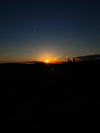A beautiful silhouette of two people sharing a moment as the sunset creates a dramatic and serene backdrop on the hill. The sky transitions into gorgeous hues.の写真素材