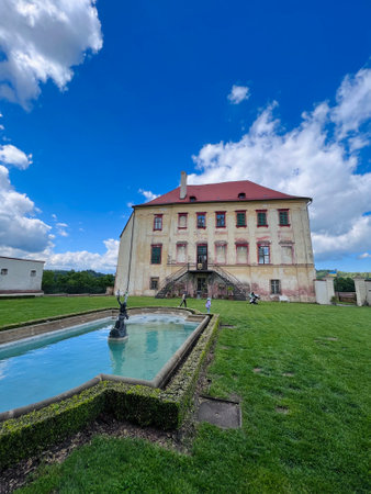 A stunning historic manor stands under a vibrant blue sky, surrounded by lush greenery in Kunstat, Czech Republic. The elegant fountain in the foreground adds charm to the serene and picturesque scene.の写真素材