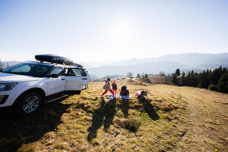 A family enjoys a peaceful picnic on a sunny day, surrounded by a beautiful mountain landscape and nature, parked with their car on a scenic hillside.の写真素材