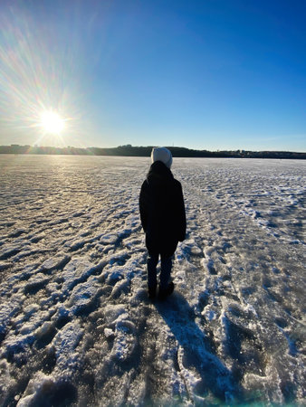 Boy stands on a frozen lake, silhouetted by the setting sun. The wintery scene evokes feelings of...