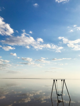 A solitary swing set stands on a vast salt flat, mirroring a serene sky filled with fluffy clouds....