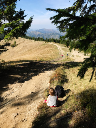 A peaceful mountain scene featuring a child sitting by a hiking trail. In the background, rolling hills and distant mountains are visible under a clear, blue sky.の写真素材