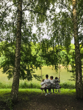 Four children sit together on a swing under tall trees by a peaceful lake, enjoying moments of carefree childhood and natureの写真素材