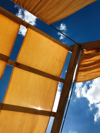 Sunlight filters through a textured fabric canopy as seen from below, framed by wooden supports against a vibrant blue sky and scattered white clouds.の写真素材