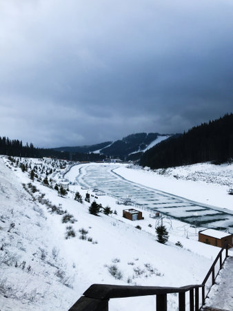A serene winter landscape featuring a frozen river winding through snow-covered hills and dense pine forests. The overcast sky adds a moody atmosphere to the peaceful scene. Bukovel,Ukraine.の写真素材