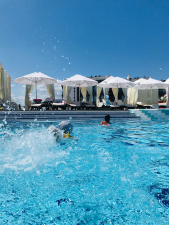 Kids playfully splash in a bright blue pool under a clear sky at a luxury resort with white...