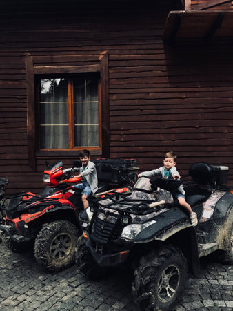 Two young boys are sitting on muddy quad bikes parked in front of a rustic wooden house, embracing...