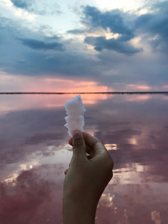 A hand holds up a salt crystal against a serene sunset over a reflective lake. The sky is painted with hues of blue, pink, and orange, creating a tranquil atmosphere.の写真素材