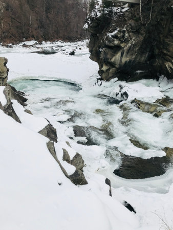 A beautiful winter scene featuring a frozen river meandering through snow-laden rocks, creating a tranquil and serene atmosphere in a woodland area.の写真素材