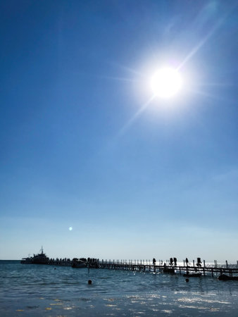 A bright sun shines over a calm ocean as people stroll along a wooden pier, creating a serene beach scene.の写真素材