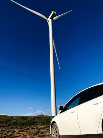 A large wind turbine towers over a white car under a clear blue sky, symbolizing renewable energy and sustainability in a natural setting.の写真素材
