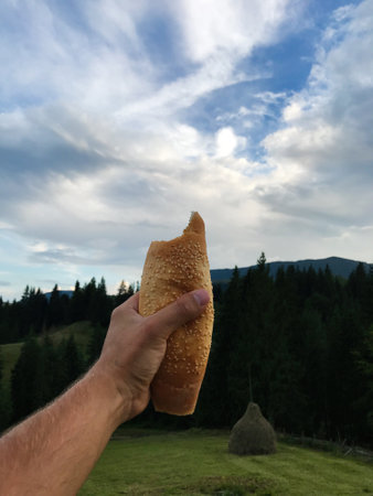 A hand holds a sesame-topped loaf of bread against a backdrop of lush mountains and a blue sky. The scene evokes a sense of tranquility and simple pleasures of life.の写真素材