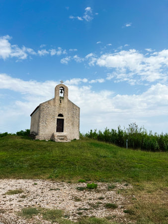 A quaint stone chapel stands on a grassy hill against a backdrop of a clear blue sky with scattered clouds, evoking a sense of peace and history. Church of Saint Vitus, Privlaka, Croatia.の写真素材