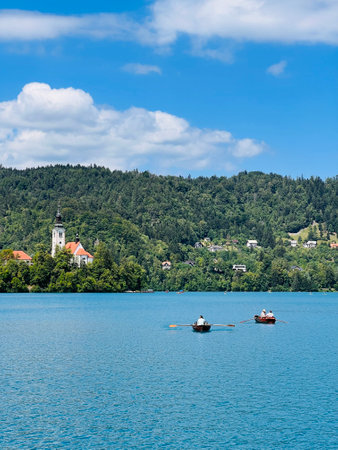 Tranquil scene of rowboats on a serene lake, surrounded by lush green mountains and a picturesque church under a vibrant blue sky with clouds. Bled, Slovenia.の写真素材