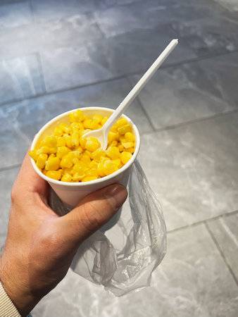 A hand holds a white disposable cup filled with bright yellow corn kernels mixed with seasoning. The...