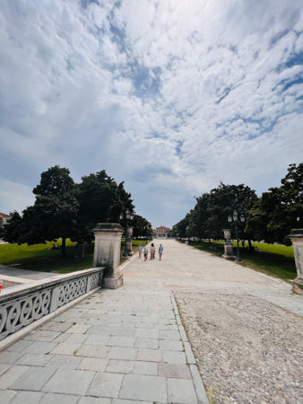 A serene park setting featuring a wide cobblestone pathway bordered by trees and grass, under a cloudy sky. People stroll along the path, enjoying the tranquil environment. Padua, Italy.の写真素材