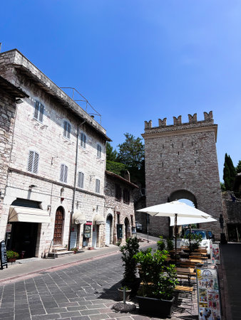 This picturesque Italian street features historic stone buildings, a quaint outdoor cafe with umbrellas, and a bright blue sky. Perfect for showcasing traditional European architecture. Assisi, Italy.の写真素材