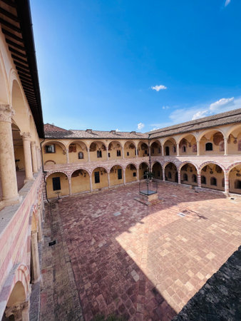 A beautiful historic courtyard surrounded by arches under a bright blue sky, showcasing classic architecture.  Assisi, Italy.の写真素材