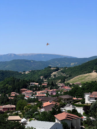 A small aircraft soars over a picturesque village surrounded by lush green hills and distant...