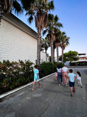A group of children and adults walk together on a quiet, palm-lined street. The atmosphere is relaxed, capturing moments of togetherness and exploration under a bright blue sky. Ippocampo, Italy.の写真素材
