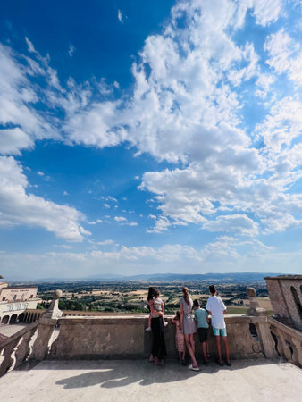 A family stands on a historic balcony, gazing at the vast scenic landscape under a vibrant blue sky with fluffy clouds, capturing a moment of togetherness and serenity. Assisi, Italy.の写真素材