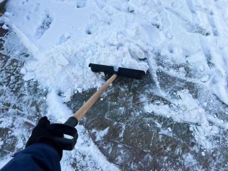A close-up view of a person's hand as they use a manual shovel to clear snow from a brick pathway, showcasing winter outdoor activity, snow removal, and cold weather maintenance.の写真素材