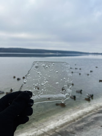 A close-up of a person holding a clear ice shard outdoors with a serene lake in the background,...