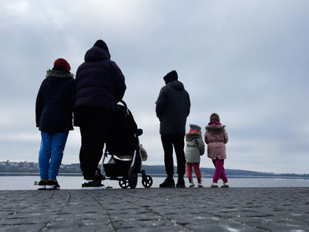 A group of children and adults stand together facing a tranquil lake under a cloudy sky.の写真素材