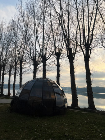 A reflective geodesic dome situated on grass beside a calm river, lined with bare trees under a...