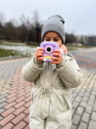 A young child wearing winter clothing is focused on using a colorful toy camera outdoors, enjoying...
